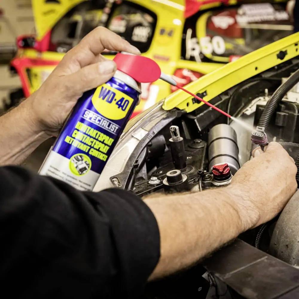 'Eine Person sprüht WD-40 auf einen Autoteilmotor in einer Werkstatt, im Hintergrund sind Rennwagen sichtbar.'