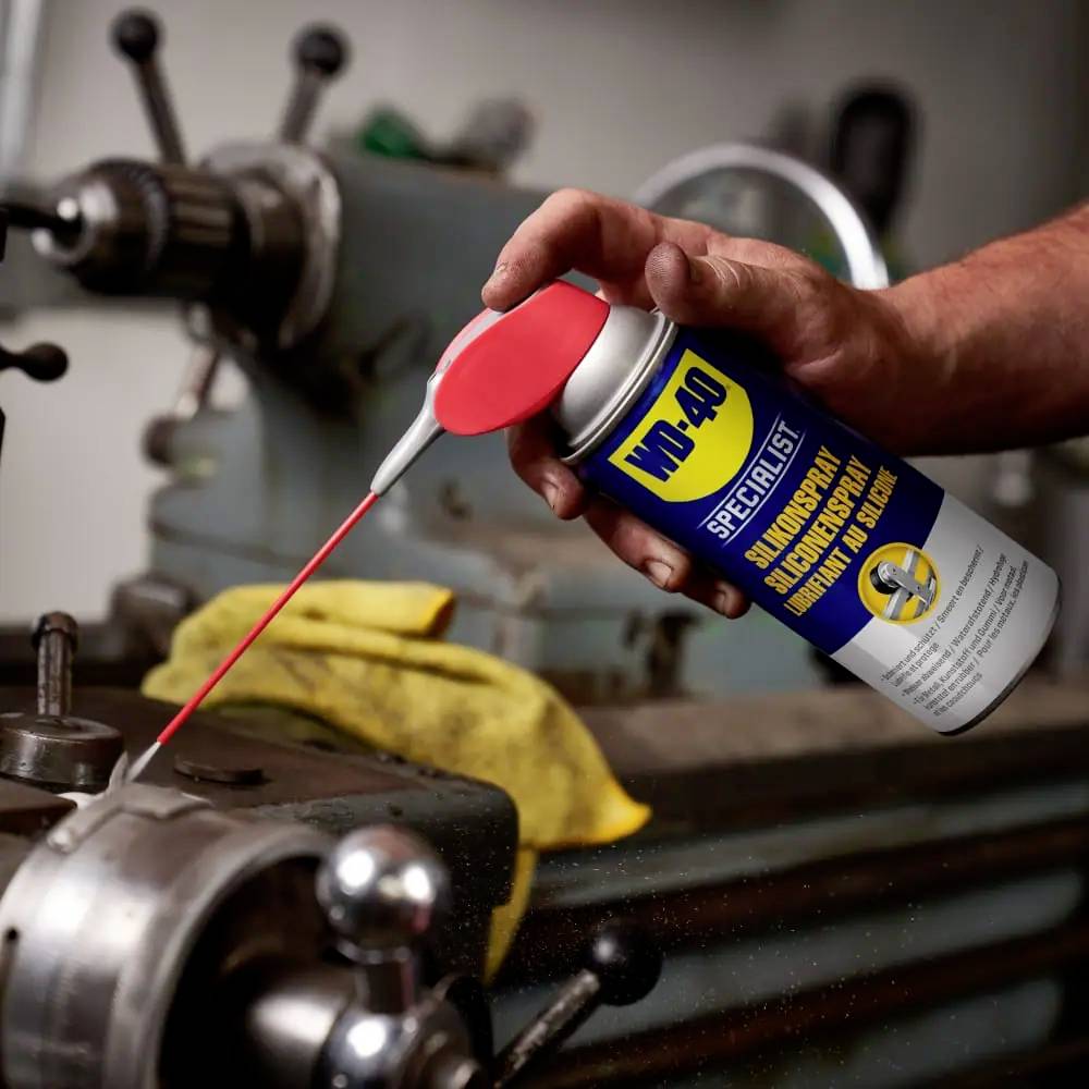 Eine Person sprüht Schmiermittel aus einer WD-40-Dose auf den beweglichen Teil einer alten Drehbank, um sie zu warten.