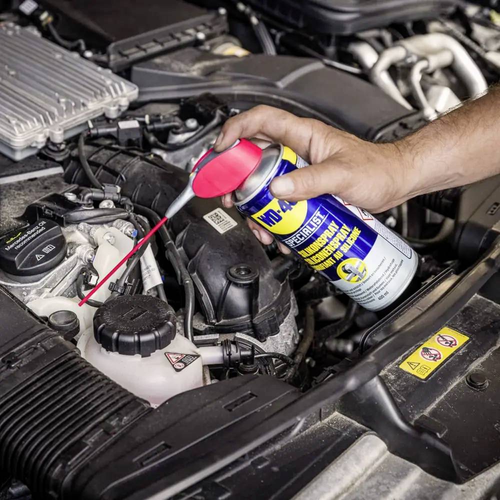 Eine Hand sprüht ein Schmiermittel auf einen Autoteilmotor, um Wartung durchzuführen. Der Motorraum ist deutlich sichtbar.