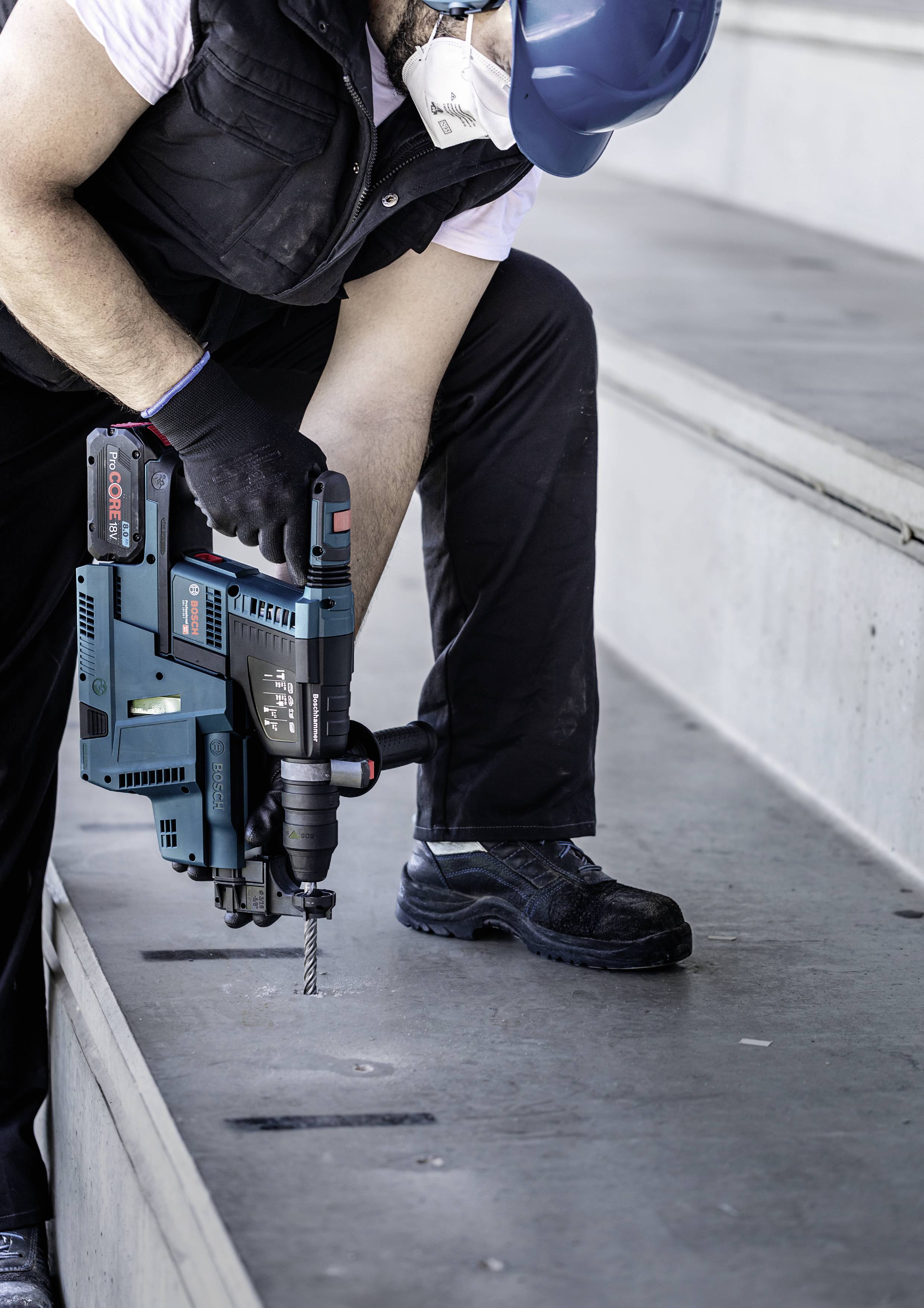 Ein Arbeiter mit blauem Helm und Schutzmaske bohrt mit einem Elektrowerkzeug in Betonstufen, trägt Handschuhe und Arbeitskleidung.