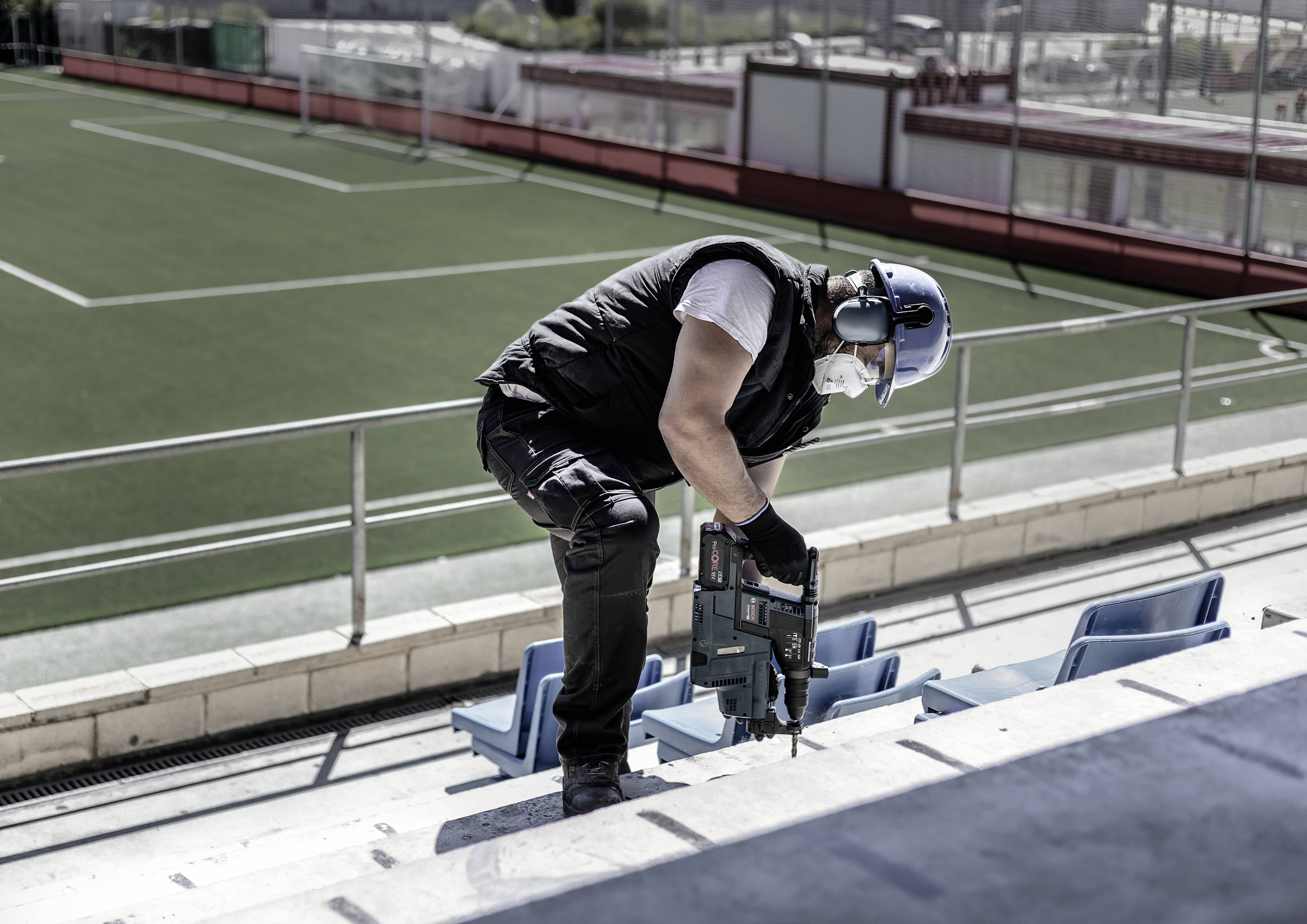 Mann arbeitet mit Elektrowerkzeug auf Stadiontribüne, trägt Helm und Gehörschutz, leere Fußballspielfläche im Hintergrund.
