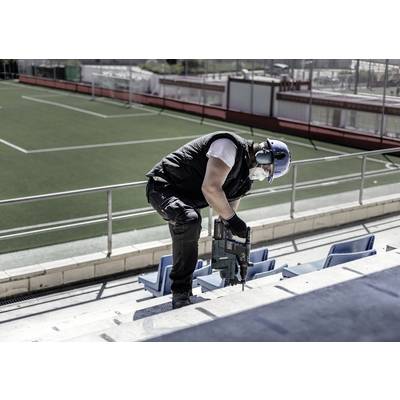 Eine Person arbeitet mit Baugeräten auf einer Stadiontribüne neben einem Fußballfeld. Sie trägt Schutzkleidung und Kopfhörer.