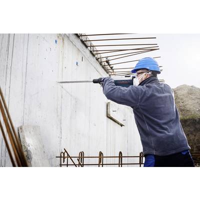 Ein Bauarbeiter in Schutzkleidung und Helm arbeitet mit einer Bohrmaschine an einer Betonmauer auf einer Baustelle.