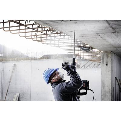Ein Bauarbeiter mit Schutzkleidung und Helm bohrt ein Loch in eine Betondecke auf einer Baustelle. Bewehrungsstahl ist sichtbar.