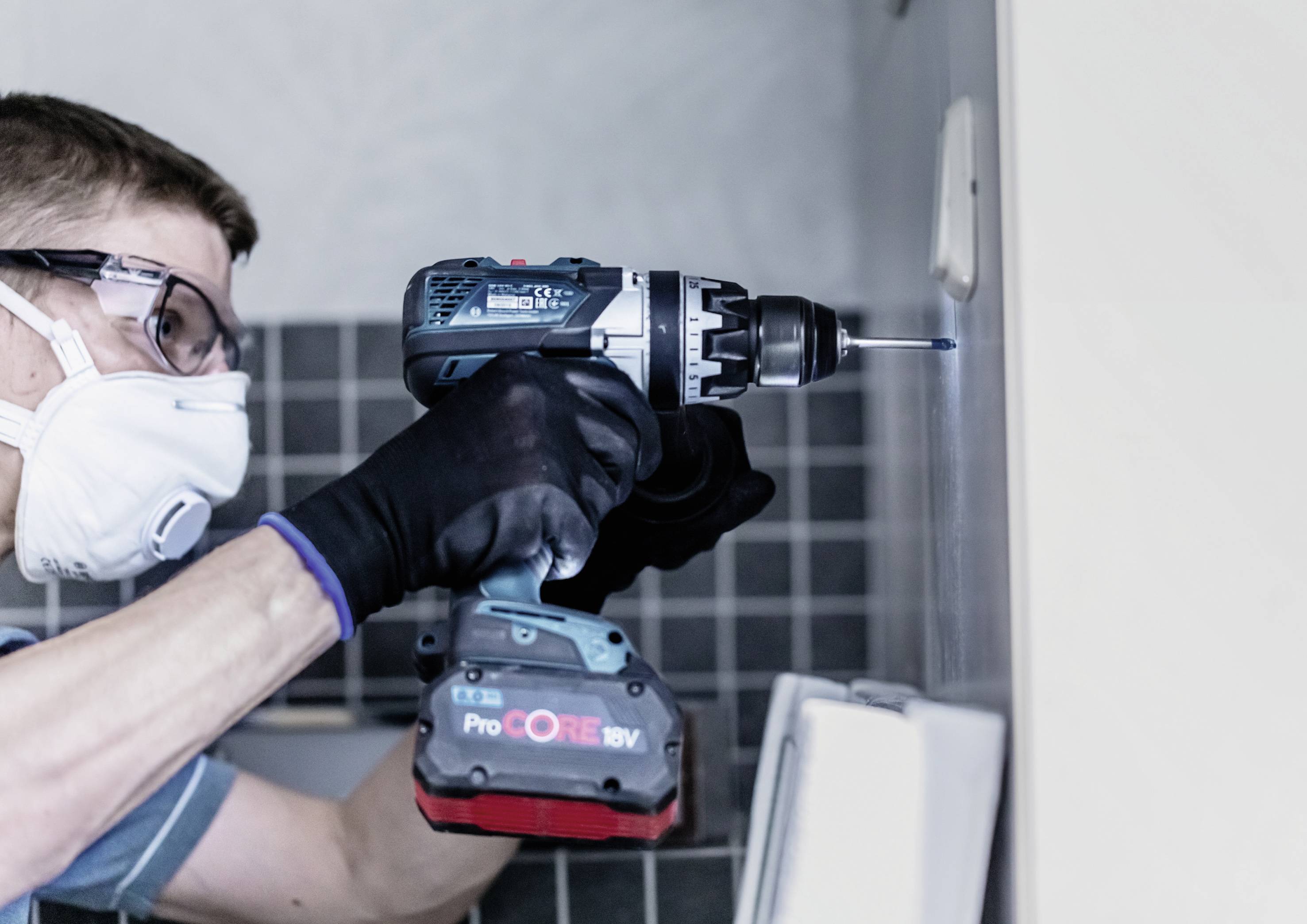 Eine Person mit Schutzbrille und Maske benutzt eine elektrische Bohrmaschine, um ein Loch in eine geflieste Wand zu bohren.