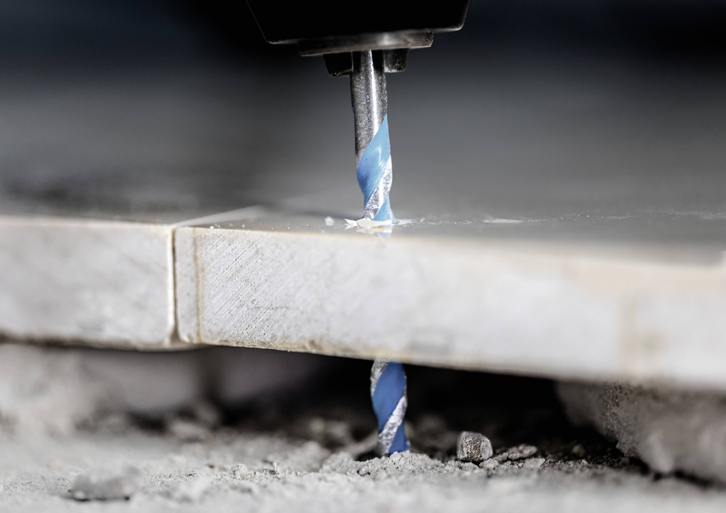 Bohrer durchdringt zwei Steinplatten auf Baustelle; Staub und Trümmer sind sichtbar.