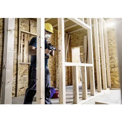 Arbeiter mit Schutzhelm und Maske benutzt Bohrer auf einer Baustelle aus Holzrahmen. Hintergrund zeigt unvollständige Wandstrukturen.