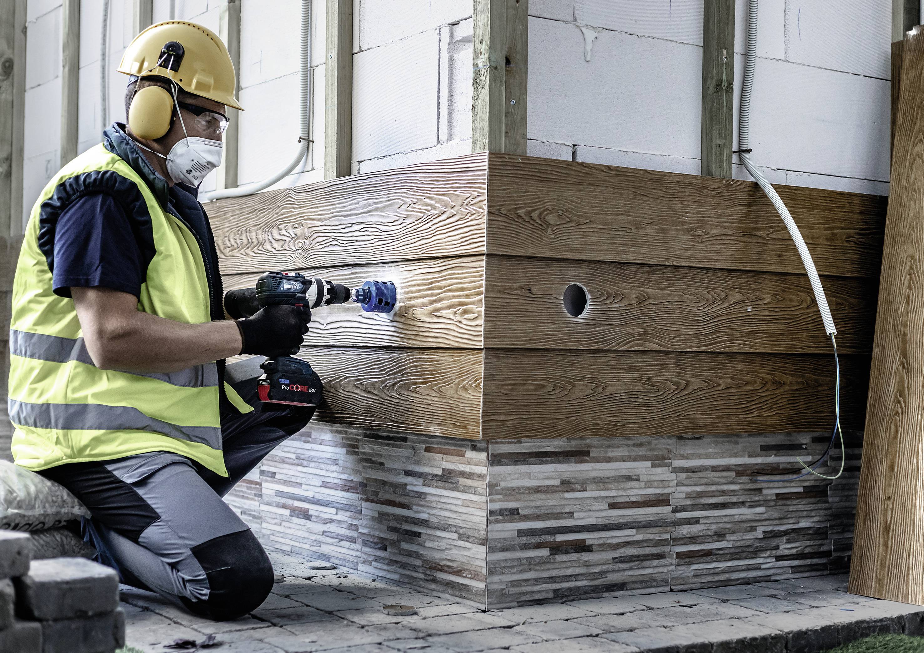 Ein Arbeiter mit Helm und Schutzbrille bohrt ein Loch in eine Holzfassade eines Gebäudes. Er trägt eine gelbe Weste und Gehörschutz.