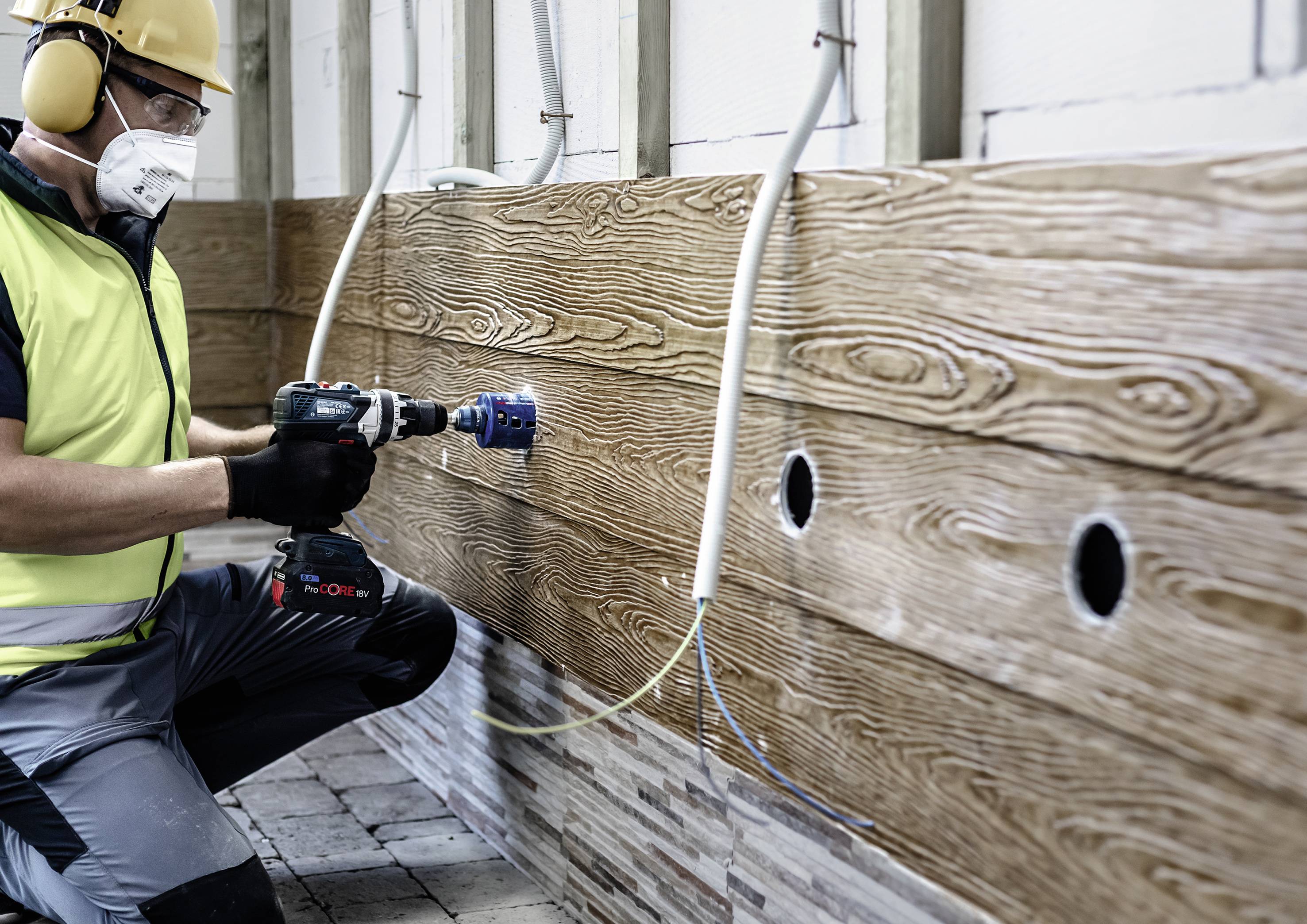 Ein Arbeiter in Schutzkleidung bohrt mit einer elektrischen Bohrmaschine Löcher in eine Wand aus Holzpaneelen.