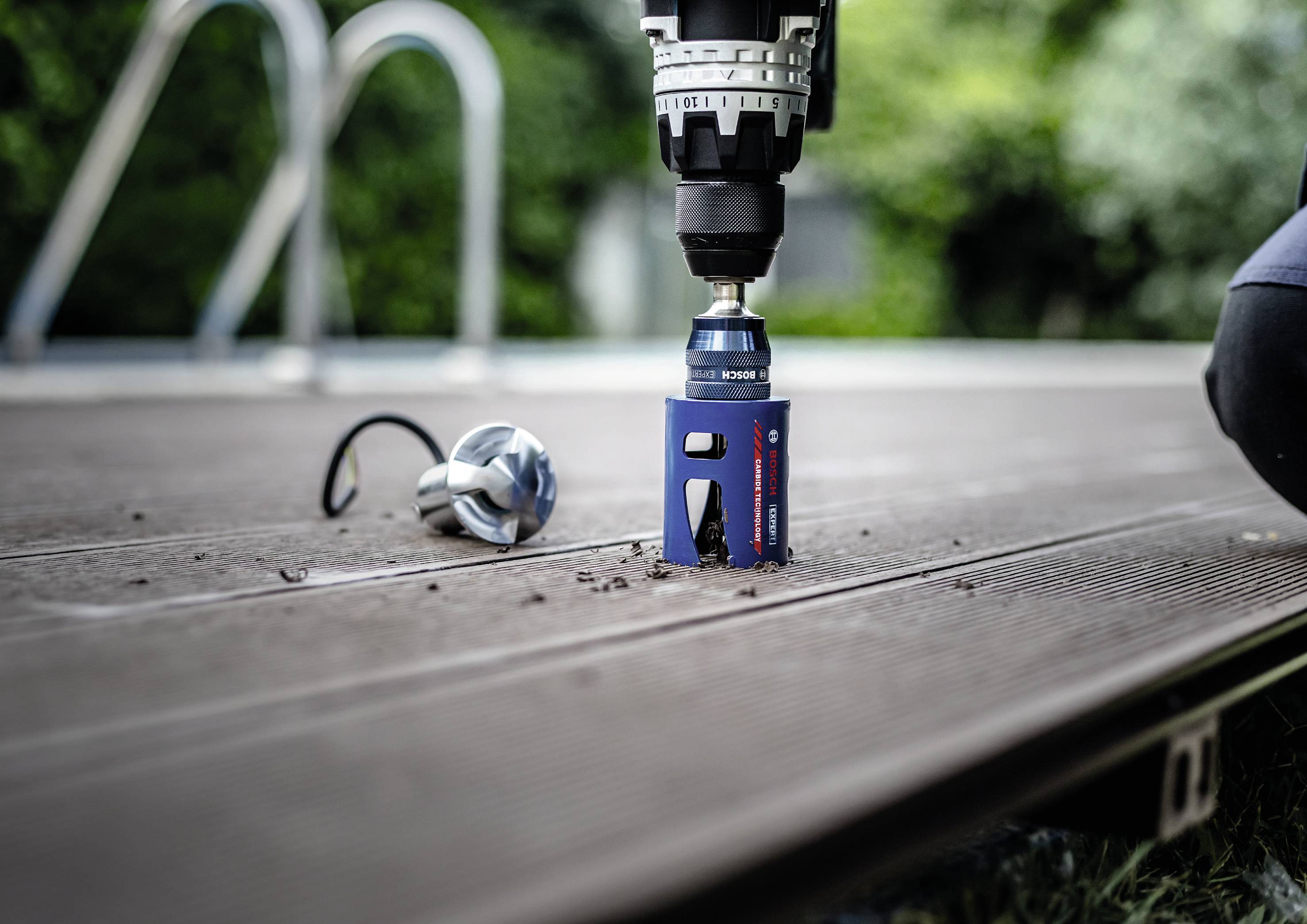 Eine Bohrmaschine bohrt ein rundes Loch in ein Holzdeck im Freien. Neben der Bohrstelle liegt ein Metallteil auf dem Deck.