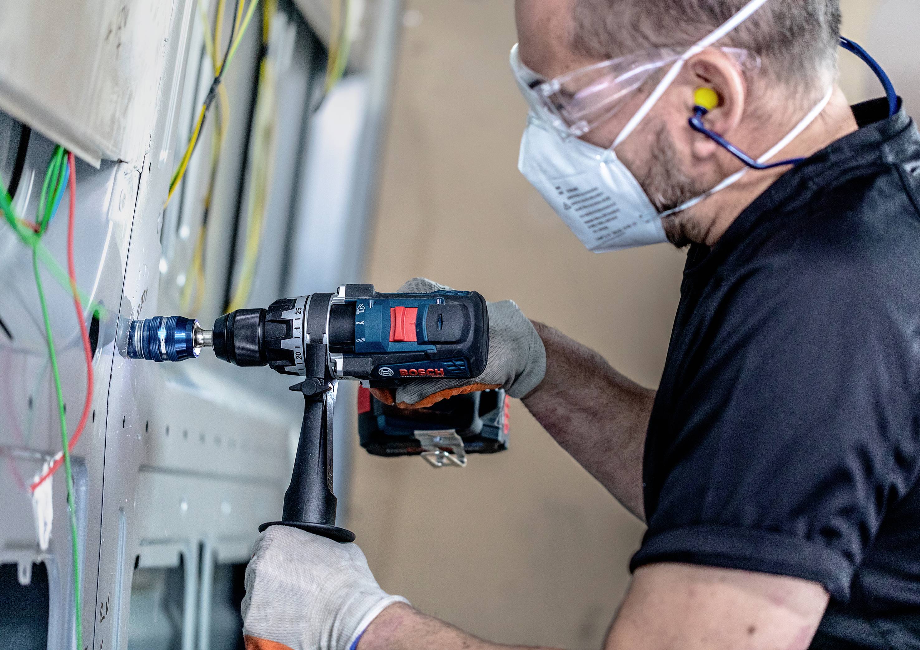 Ein Mann in Sicherheitsbrille und Maske bohrt mit einer elektrischen Bohrmaschine ein Loch in die Wand, umgeben von Drähten.