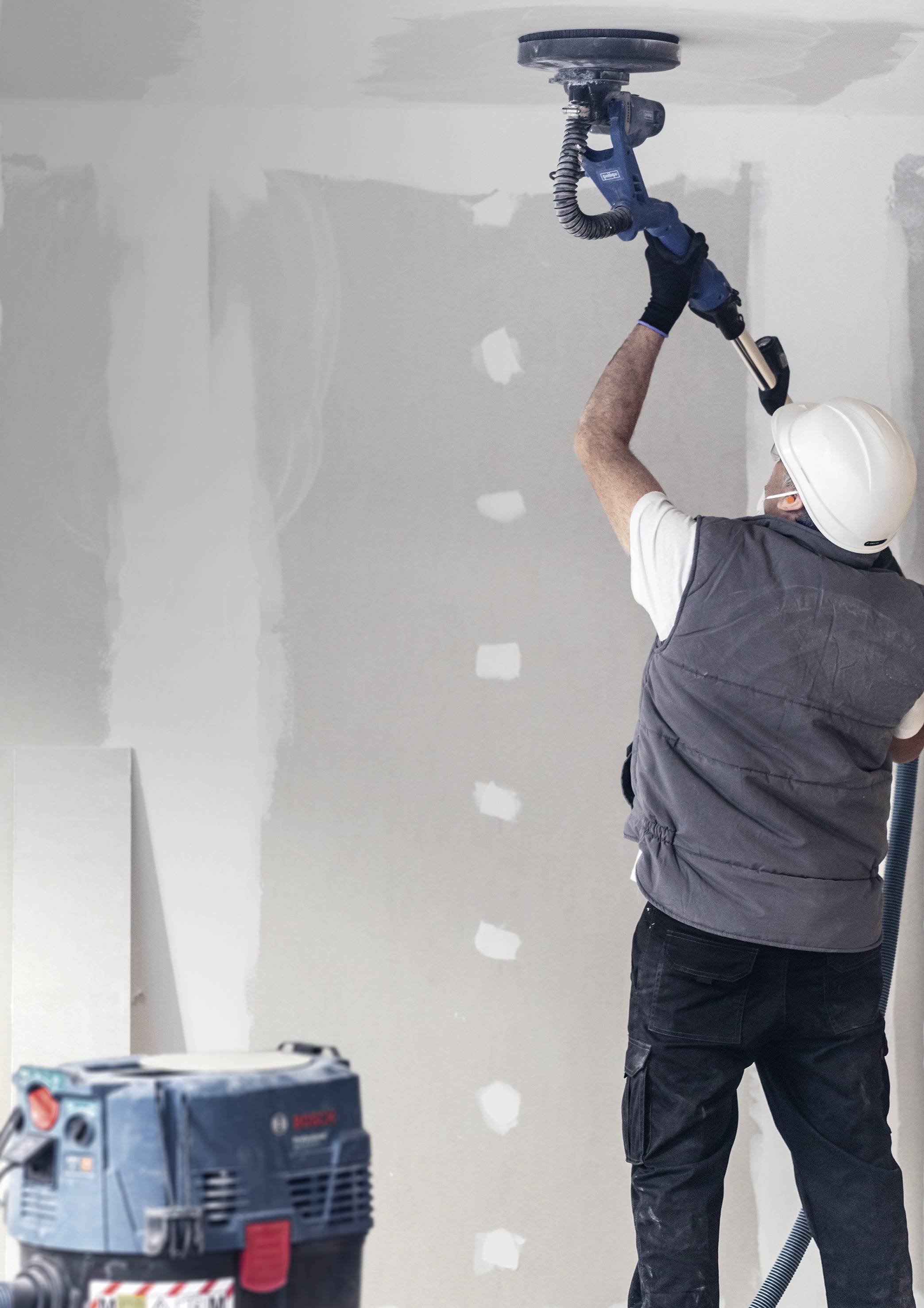 Ein Arbeiter mit Schutzhelm schleift eine Gipskartonwand, während ein Schleifgerät den Staub absaugt. Im Hintergrund sind weiße Markierungen auf der Wand sichtbar.