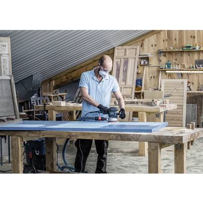 Ein Mann arbeitet in einer Werkstatt, trägt Schutzbrille und schleift eine Holzplatte auf einem Tisch. Im Hintergrund sind Werkzeuge sichtbar.