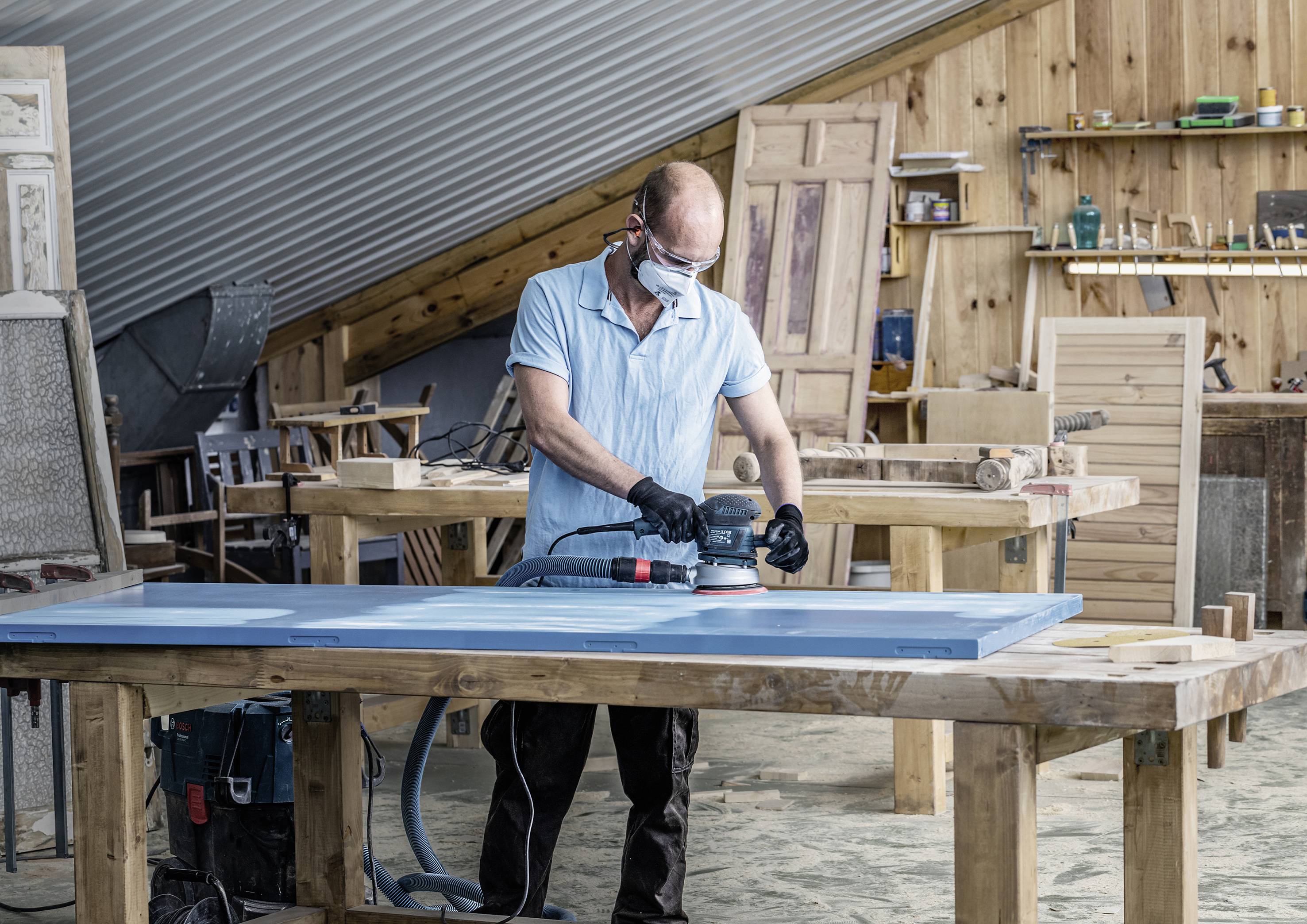Ein Mann arbeitet in einer Werkstatt und schleift mit einer Maschine eine Holzplatte. Umgeben von Werkzeugen, trägt er Schutzbrille und Ohrschützer.