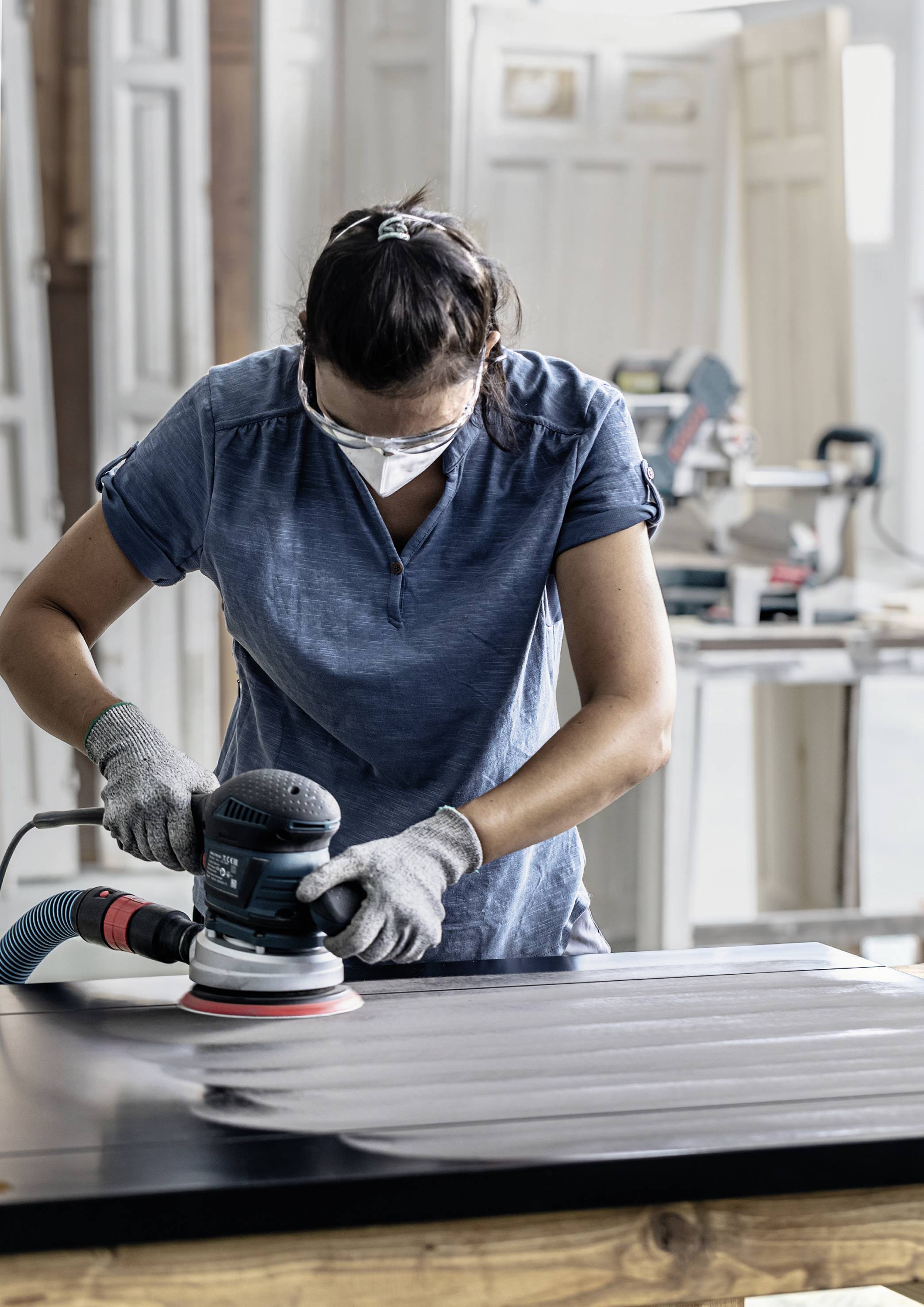 Person beim Schleifen einer Holzplatte mit einer elektrischen Schleifmaschine, trägt Schutzbrille und Handschuhe, in einer Werkstatt.