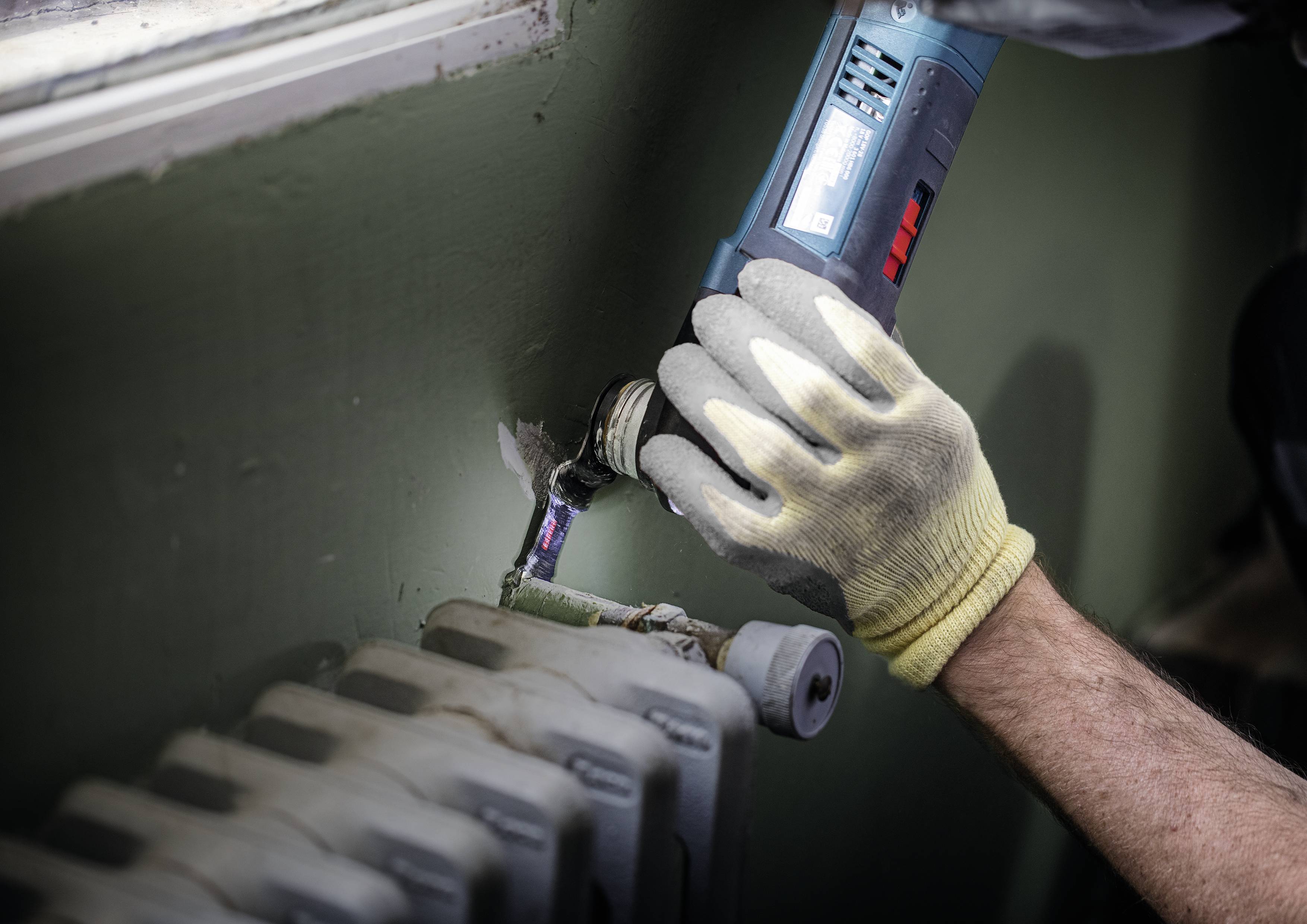 Ein Handwerker in gelben Handschuhen arbeitet mit einem Elektrowerkzeug an einem Heizkörperventil, um Reparatur- oder Wartungsarbeiten durchzuführen.