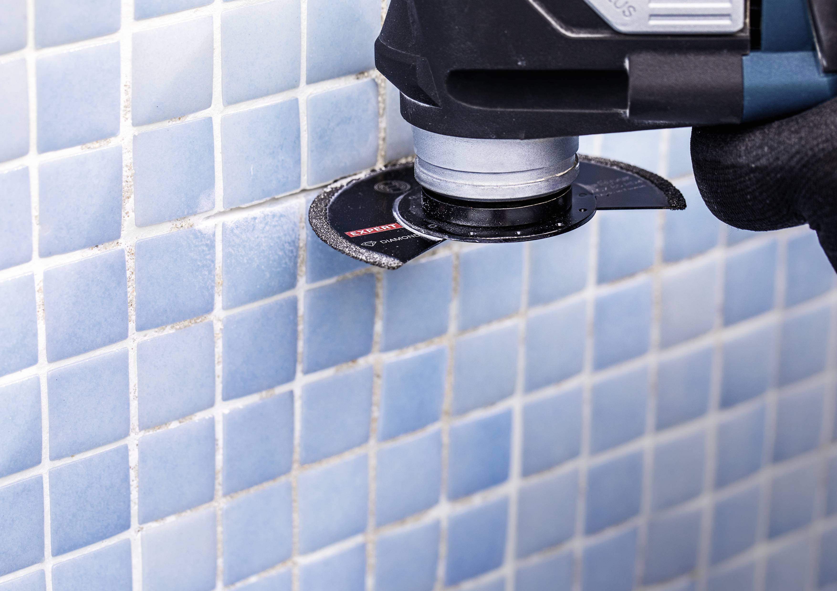 Eine Elektrosäge schneidet Mosaikfliesen an einer Wand. Die Fliesen sind blau und quadratisch angeordnet.