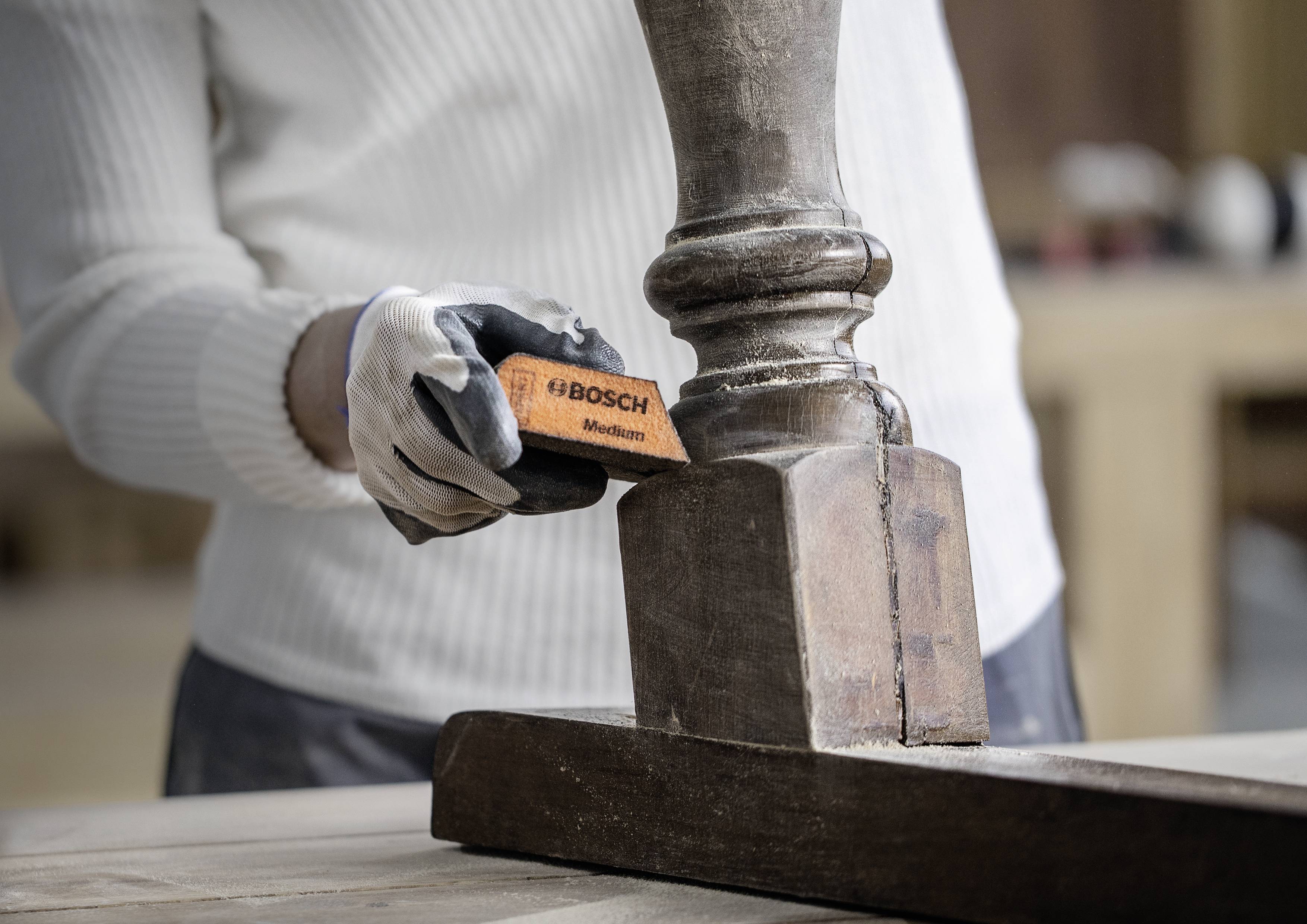 Person schleift einen Holzpfosten mit einem orangen Schleifklotz. Der Fokus liegt auf der präzisen Handarbeit.