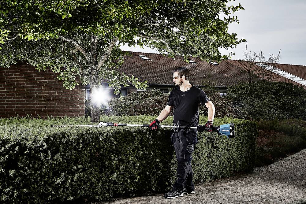 Ein Mann schneidet mit einer Heckenschere eine Hecke. Im Hintergrund ist ein Backsteingebäude und ein großer Baum zu sehen.