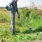 Eine Person in Schutzkleidung schneidet Gras mit einem elektrischen Freischneider auf einer Grünfläche. Im Hintergrund Gebäude.