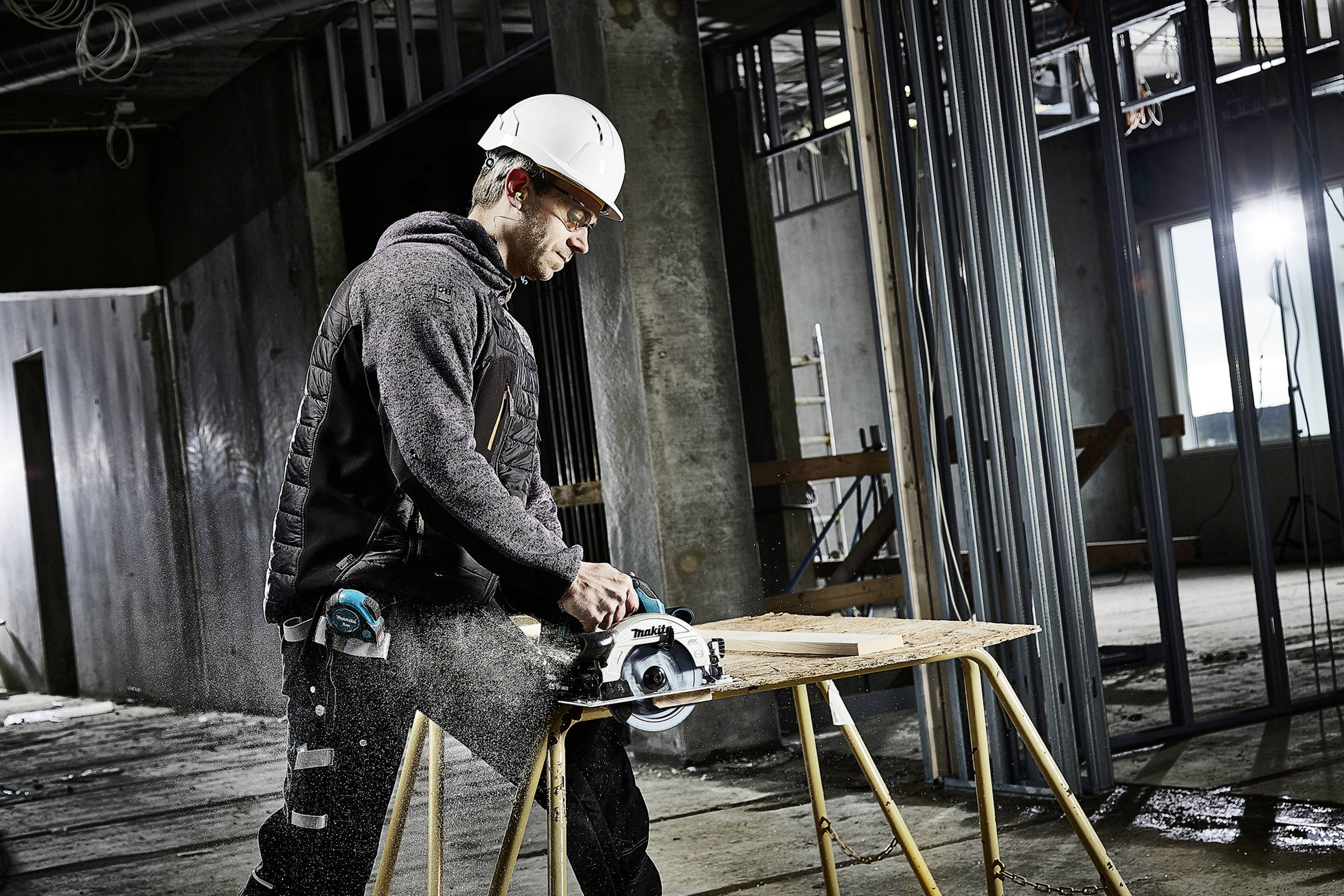 Ein Bauarbeiter mit Schutzhelm schneidet Holz mit einer Kreissäge in einem unfertigen Gebäude. Umgebend sind Metallrahmen sichtbar.
