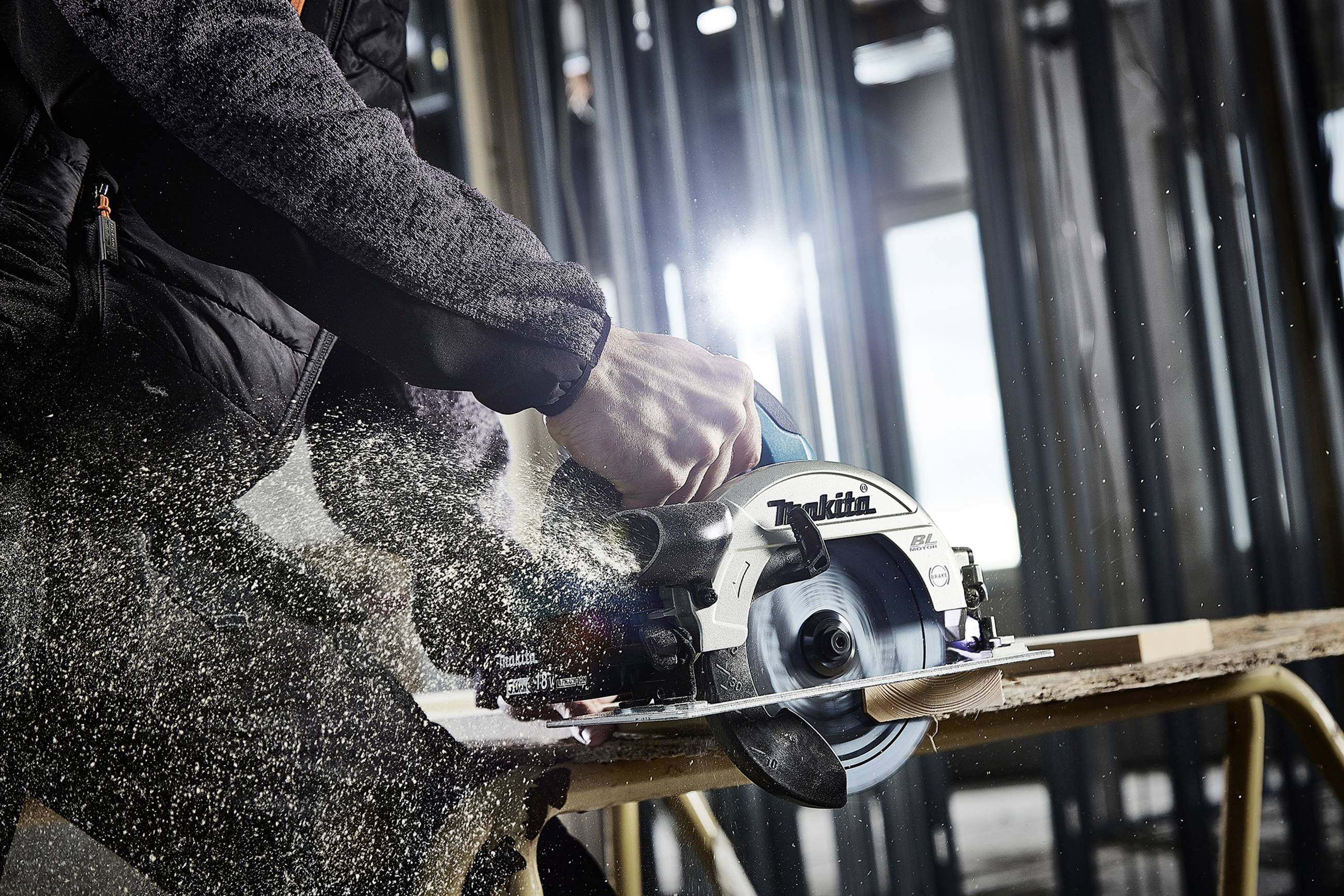 Ein Handwerker sägt mit einer elektrischen Kreissäge ein Holzbrett, während Sägespäne wegfliegen. Im Hintergrund unscharf eine Baustelle.