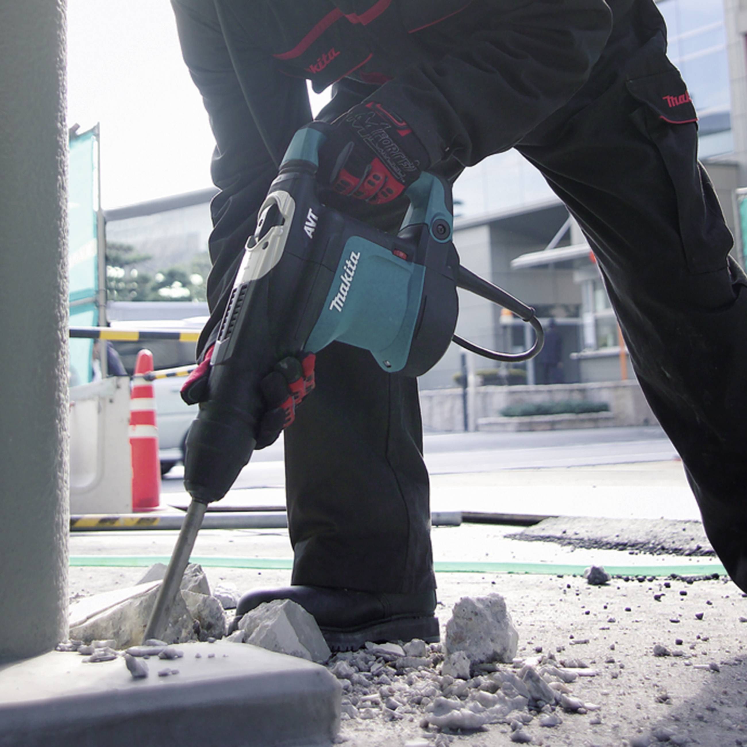 Ein Bauarbeiter in Schutzkleidung benutzt einen Bohrhammer, um Beton auf einer Baustelle zu bearbeiten. Im Hintergrund sind Gebäude zu sehen.