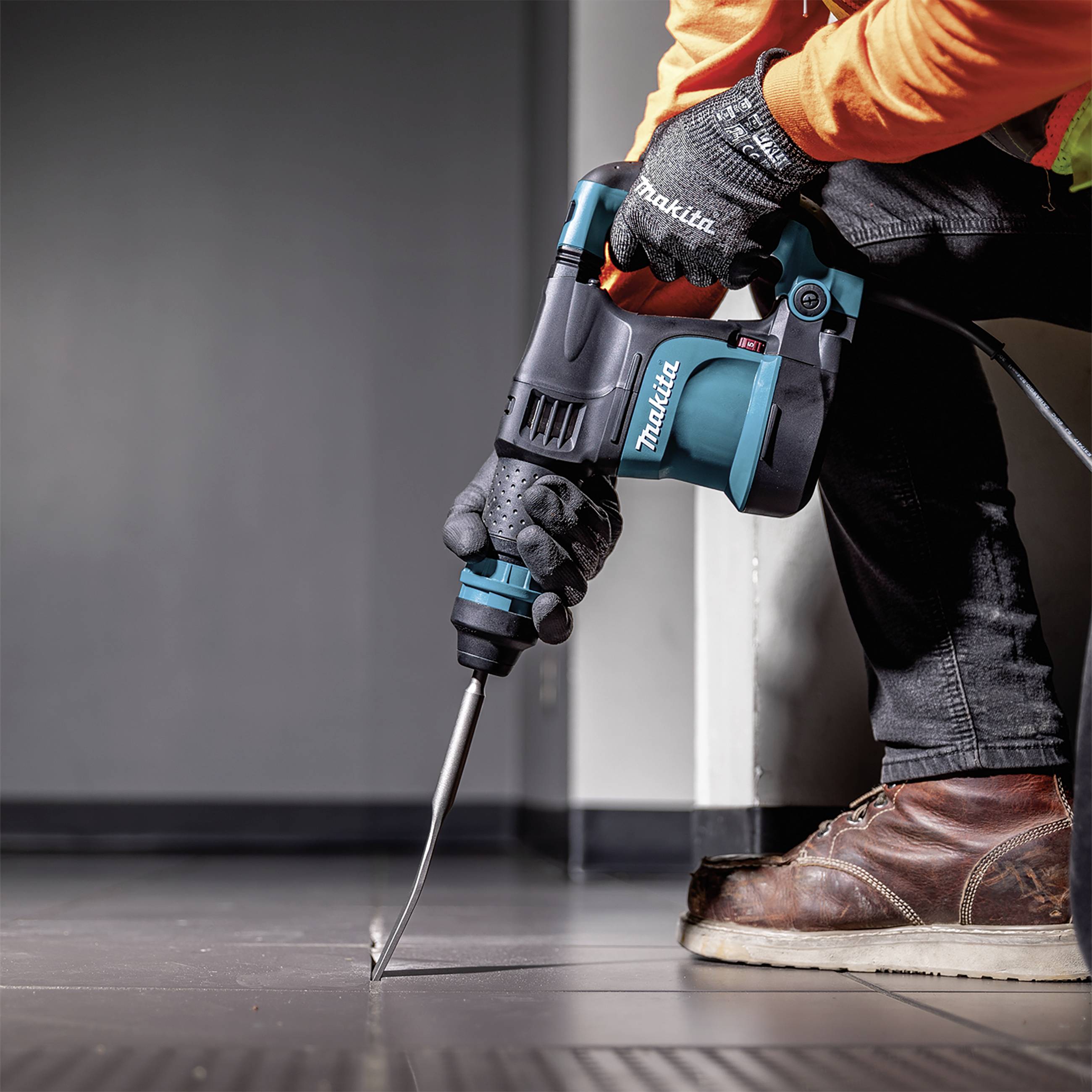 Eine Person trägt Arbeitshandschuhe und benutzt einen blauen Makita-Bohrhammer, um Fliesen auf einem Boden zu bohren oder zu meißeln.