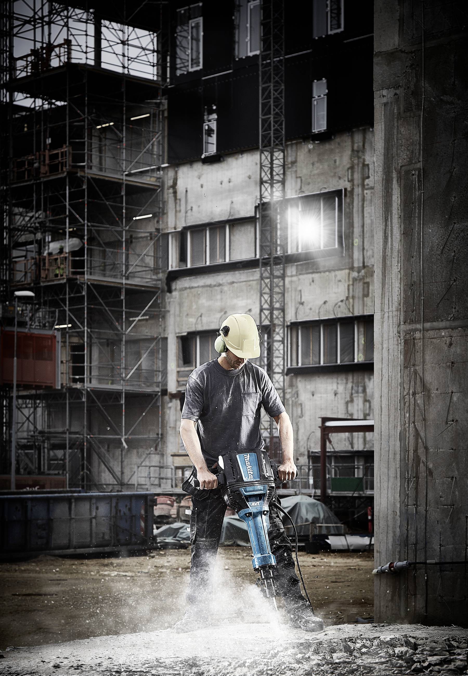 Arbeiter mit Schutzhelm bedient einen Presslufthammer auf einer Baustelle, umgeben von Gebäudegerüsten.