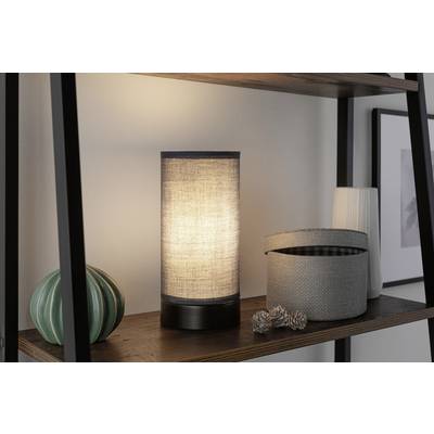 Auf einem Regal steht eine leuchtende Tischlampe mit Stoffschirm, daneben Dekorationen wie ein grüner Keramik-Kaktus und eine geöffnete Stoffbox.