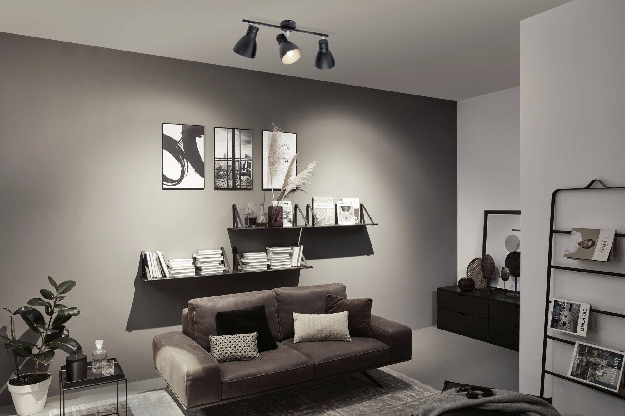 Modernes Wohnzimmer mit dunkelbraunem Sofa, dekorativen Kissen, Wandbildern und Regalen voller Bücher. Dezente Wandbeleuchtung.