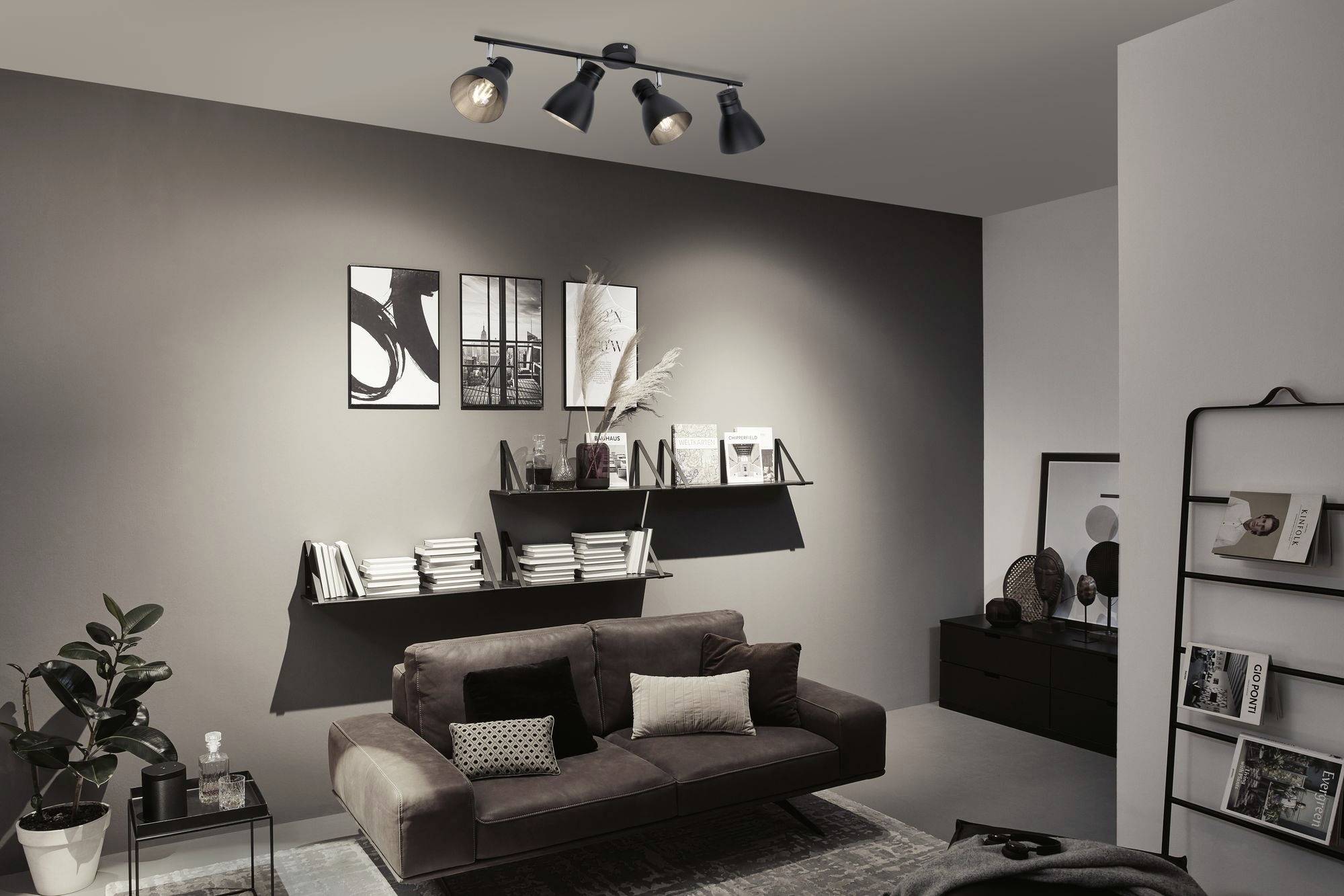 Ein modernes Wohnzimmer mit braunem Sofa, dekorativen Kissen und schwarzem Deckenstrahler. Wandregale mit Büchern und Bildern.