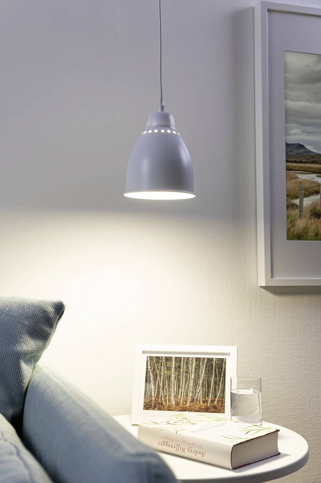 Eine Hängelampe beleuchtet ein weißes Beistelltischchen mit einem gerahmten Foto, einem Buch und einem Glas Wasser. Ein Sofa steht daneben.