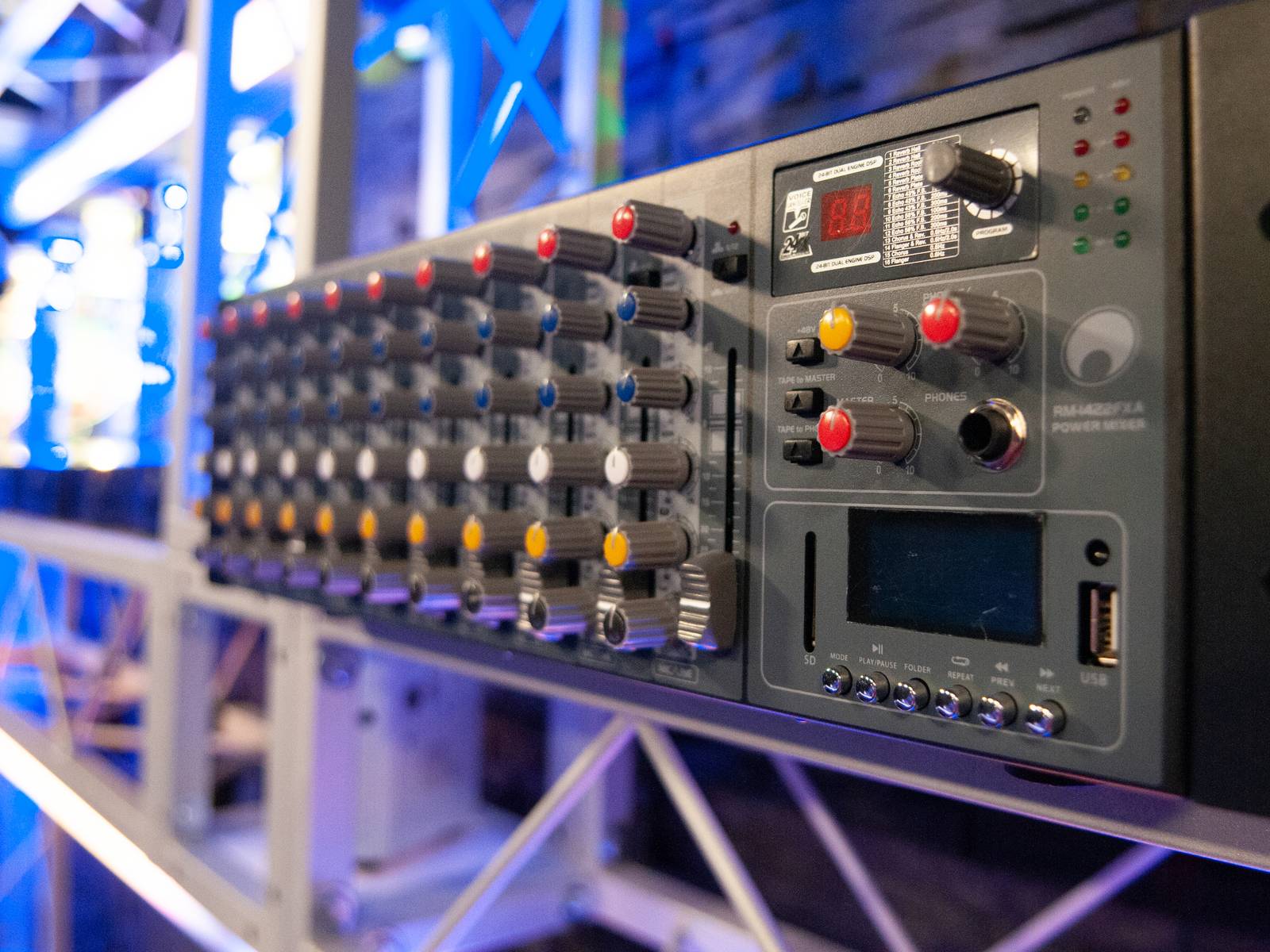 Ein modernes Audiomischpult mit vielen Reglern. Es ist Teil einer Bühnenausstattung, beleuchtet in Blau und Rosa.