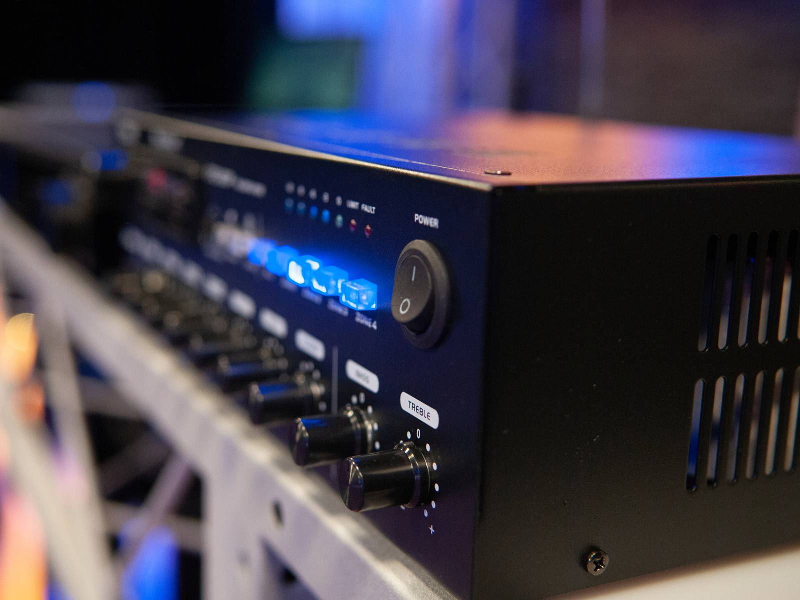 Ein schwarzer Audioverstärker mit blauen LED-Leuchten und zwei Drehknöpfen an der Vorderseite, platziert auf einem Metallregal.
