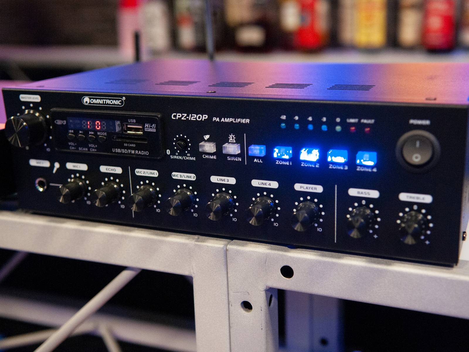 Ein schwarzer Verstärker mit mehreren Knöpfen und leuchtenden LED-Anzeigen, steht auf einem Metallgestell in einer technologisch anmutenden Umgebung.