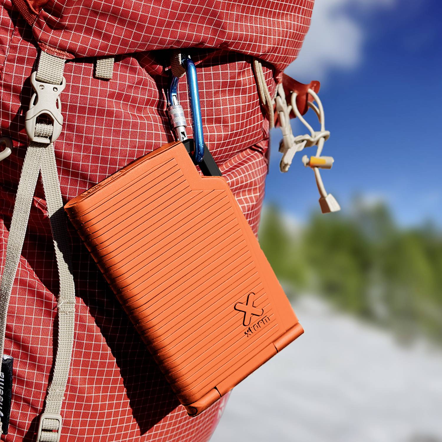 Oranger Rucksack hängt an einem Karabiner an einem größeren, roten Rucksack. Hintergrund zeigt unscharfe Natur. Outdoor-Ausrüstung.