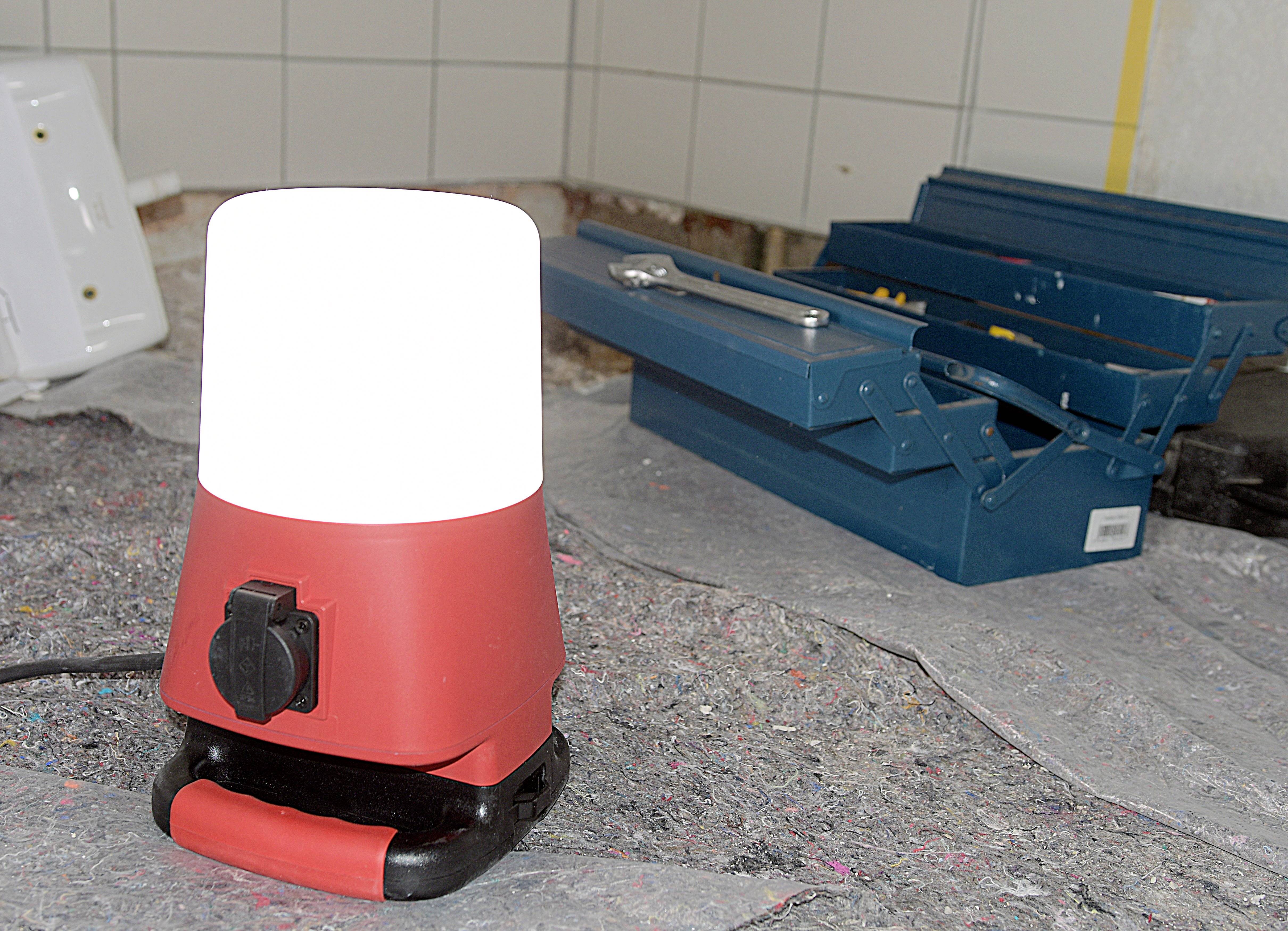 Eine rote Arbeitsleuchte leuchtet in einer Werkstatt. Neben der Lampe liegt ein blauer Werkzeugkasten auf einem Teppich.