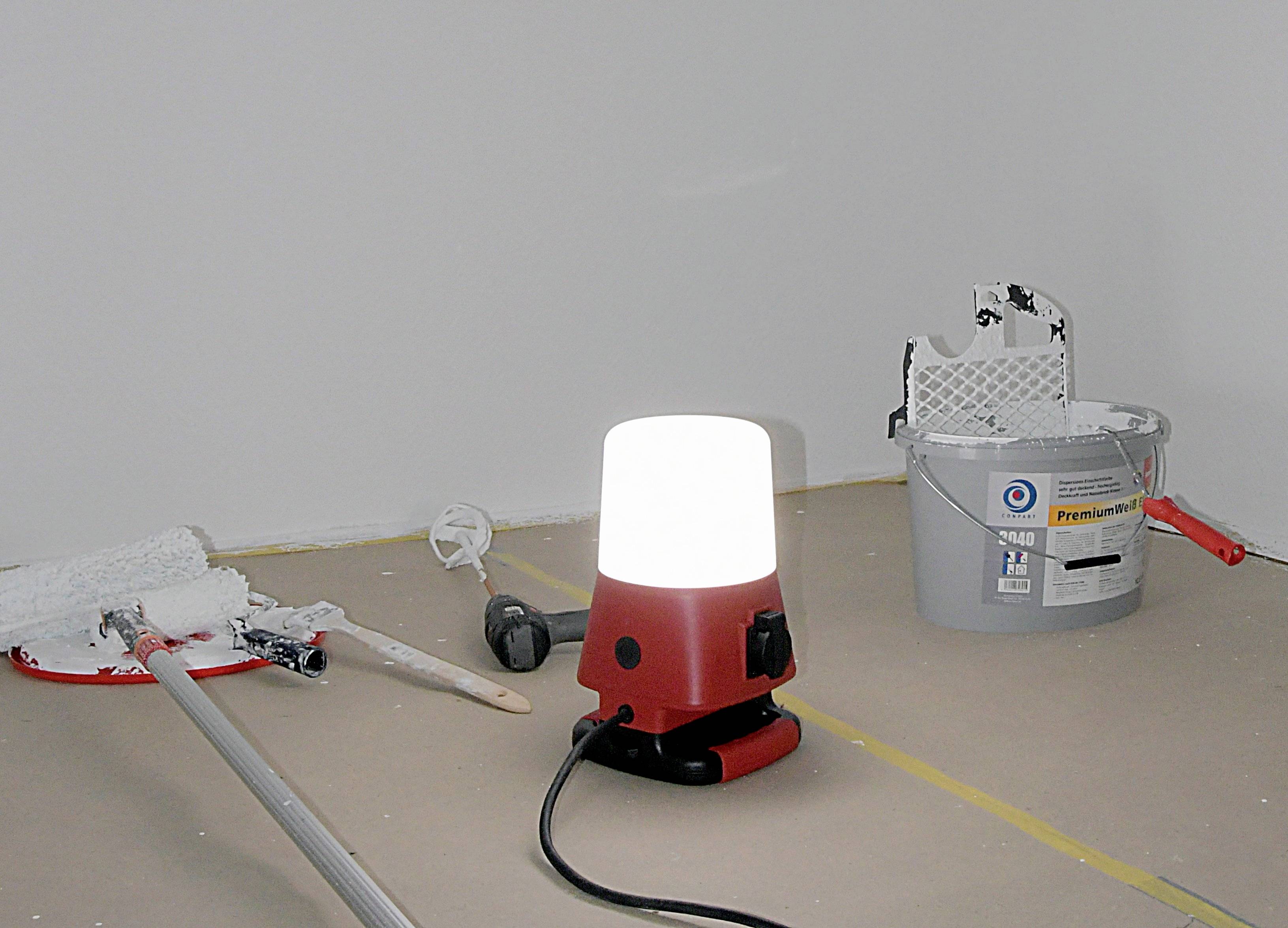 Ein Raum wird renoviert: Boden ist mit Malerschutzpapier bedeckt, Malerrolle und -eimer mit weißer Farbe sowie eine Arbeitslampe sind sichtbar.