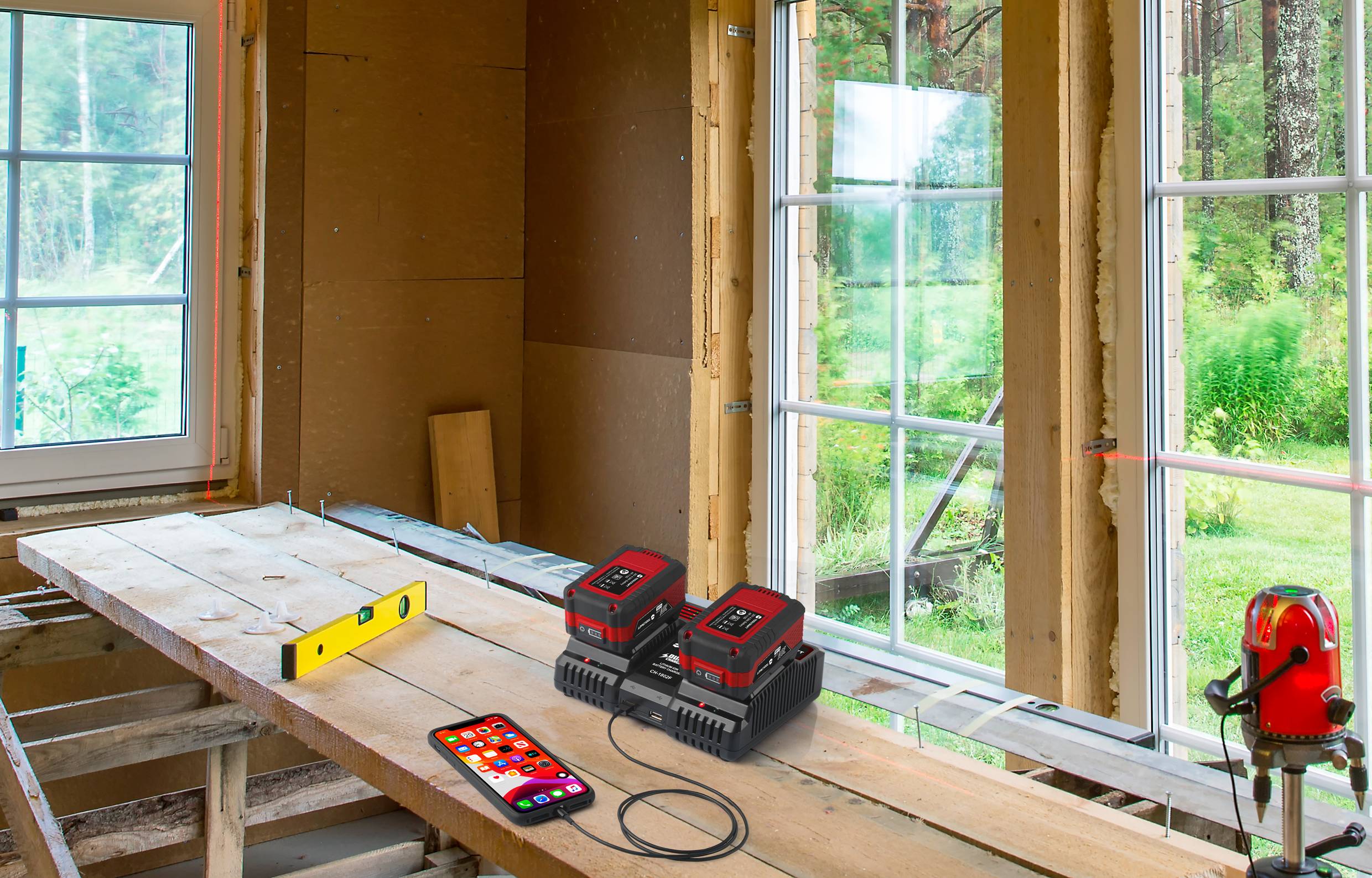 Auf einer Baustelle stehen zwei rote Akkuladegeräte mit Akkus auf einem Holztisch, daneben ein Smartphone und eine Wasserwaage.