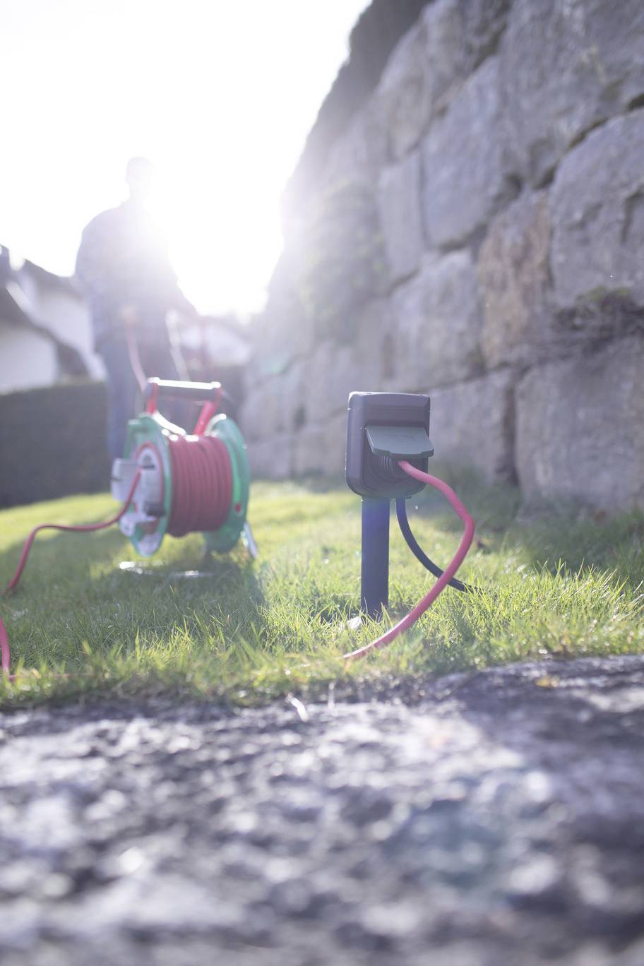 Eine Kabeltrommel im Garten, an eine Steckdose angeschlossen. Im Hintergrund unscharf eine Person neben einer Steinmauer.