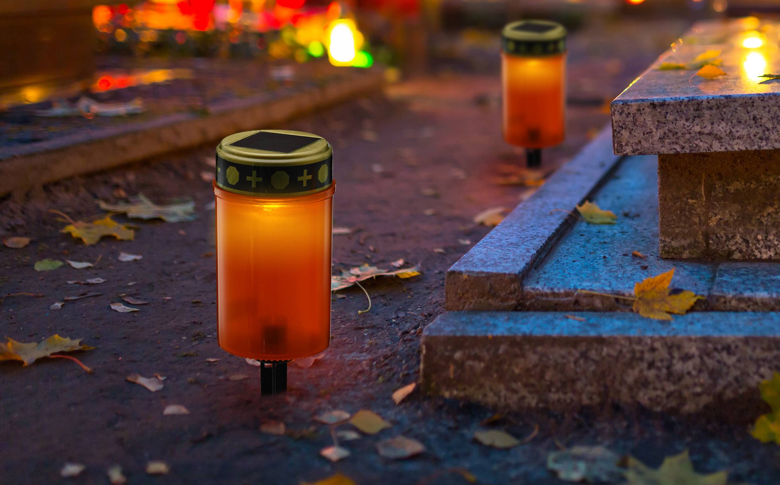 Kerzen mit Solarpanelen leuchten auf einem Friedhof in der Dämmerung. Herbstblätter liegen auf dem Boden um die Steinplatten.