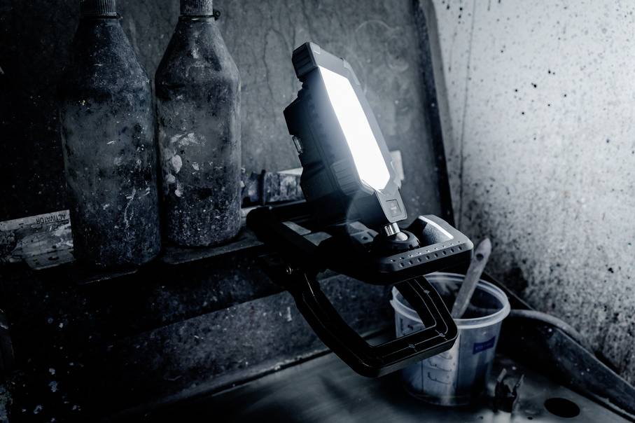 Eine leuchtende Arbeitsleuchte befindet sich in einer Werkstatt. Neben ihr stehen Flaschen mit Farbe und ein Pinsel in einem Eimer.