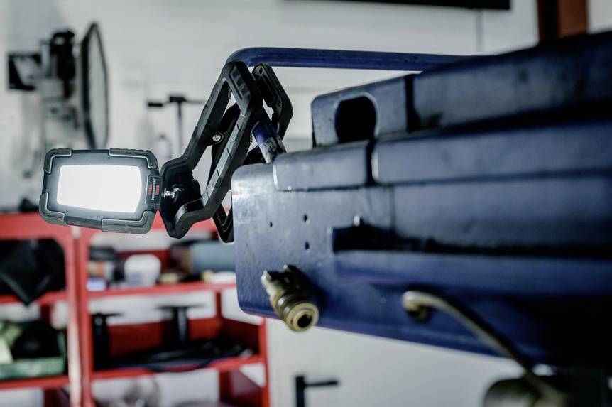 Eine helle Lampe ist an einem blauen Werkstück befestigt, im Hintergrund Regale mit Werkstattutensilien.