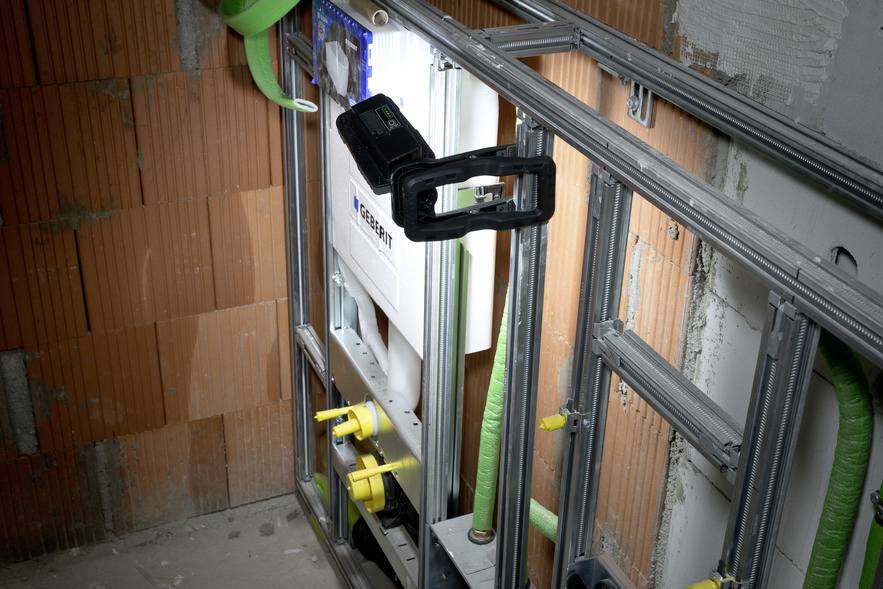 Ein Badezimmer im Bau mit sichtbaren Ziegel- und Metallstrukturen. Ein Werkzeug ist an der Wand befestigt, Rohre sind zu sehen.