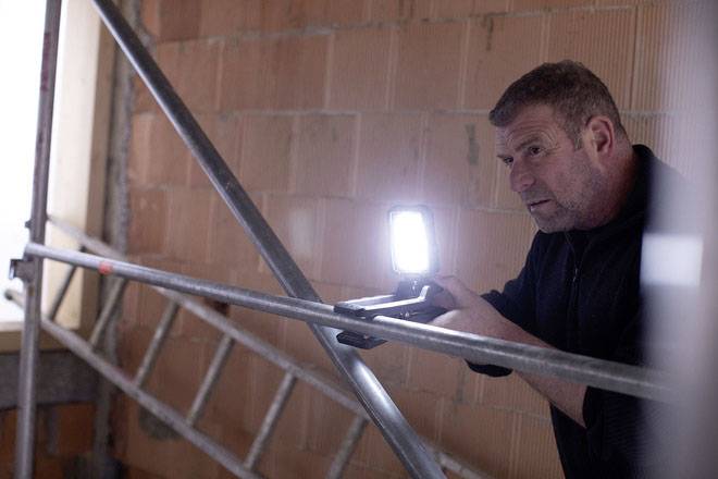 Ein Mann hält eine Lampe, während er in einem rohgebauten Raum neben einem Gerüst arbeitet. Lichtquelle betont seine Konzentration.