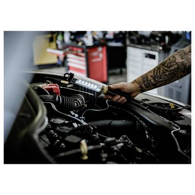 Tätowierter Mechaniker nutzt eine tragbare Arbeitsleuchte, um einen offenen Automotor in einer Werkstatt zu inspizieren.