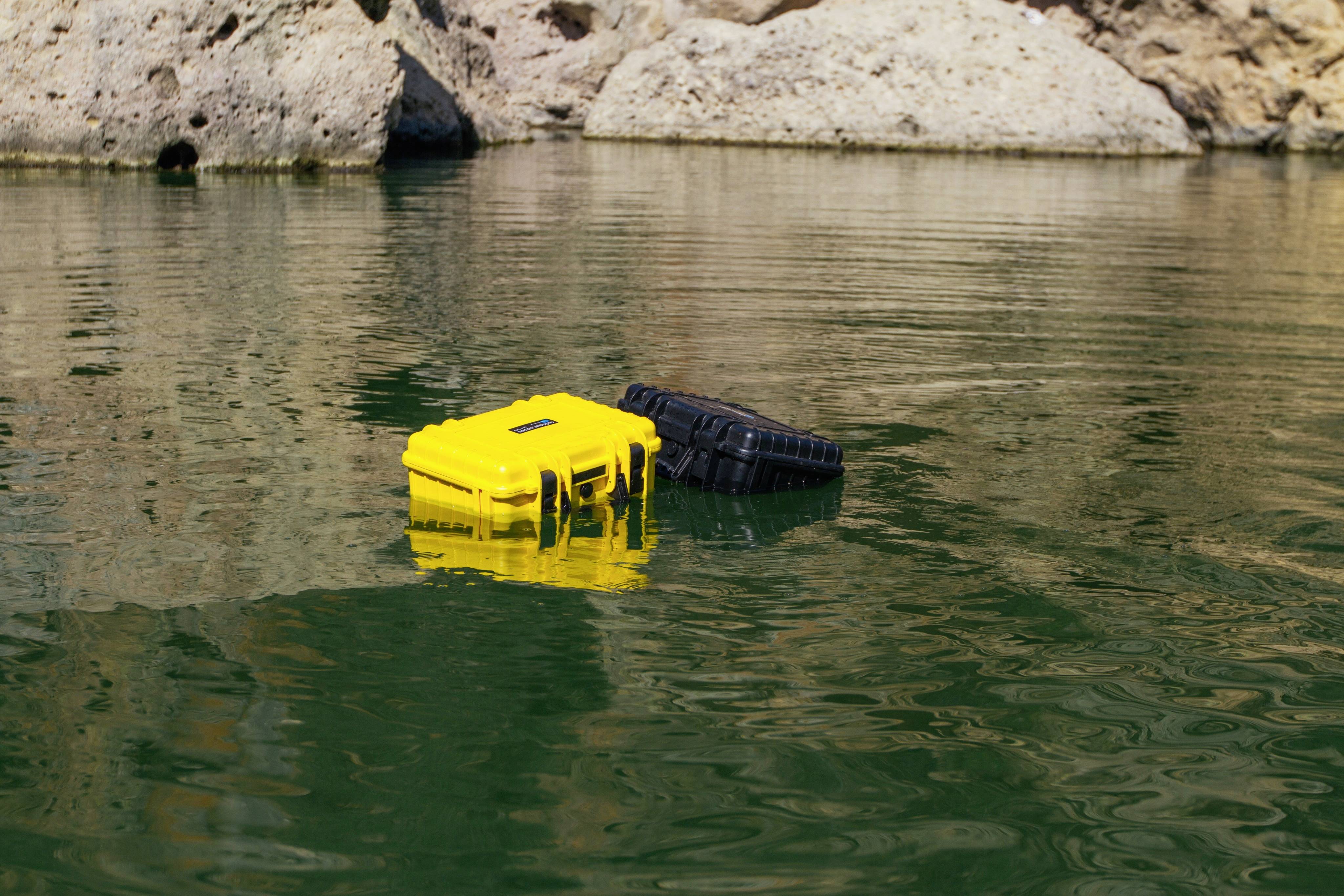 Zwei schwimmende, wasserdichte Boxen, eine gelb und eine schwarz, treiben auf grünem Wasser vor felsigem Hintergrund.