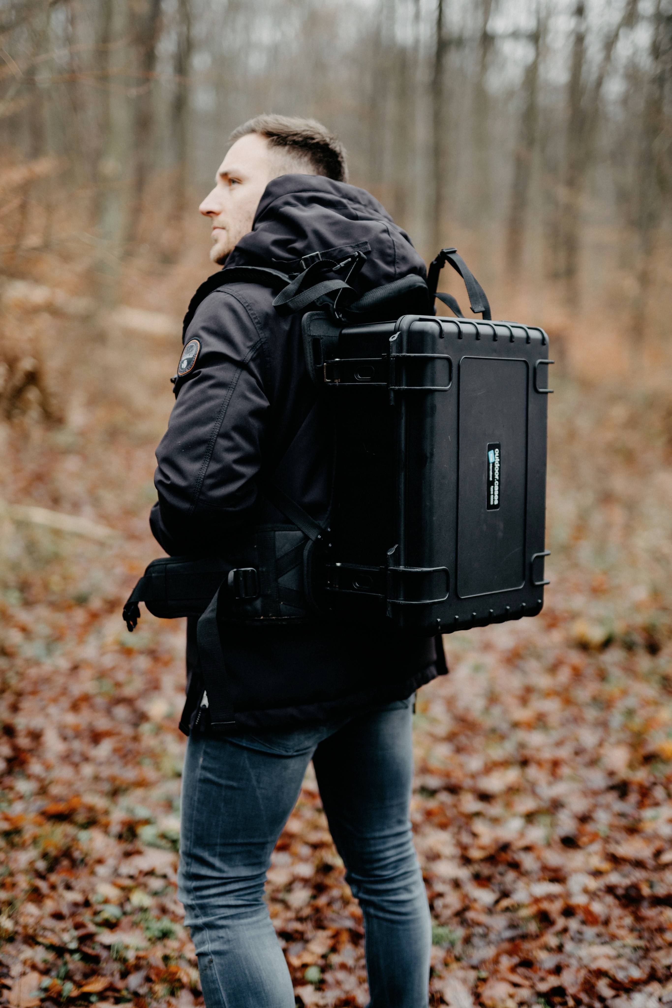 Eine Person trägt einen großen schwarzen Rucksack in einem herbstlichen Wald. Die Person trägt eine schwarze Jacke und Jeans.