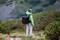 Eine Person in grüner Jacke mit großem Rucksack steht in einer bergigen Landschaft und schaut in die Ferne, umgeben von grünen Büschen.